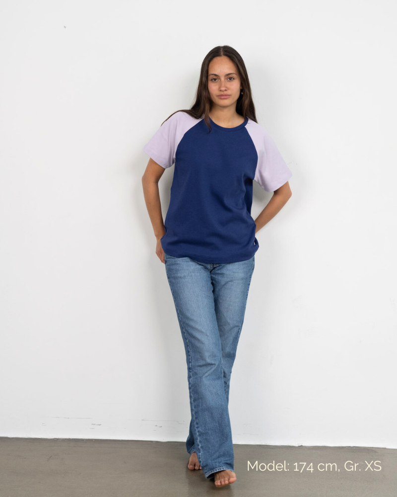 Eine Frau trägt das Raglan Shirt von Oktopulli in der Farbe Blau mit fliederfarbenen Ärmeln und lehnt dabei an einer Wand